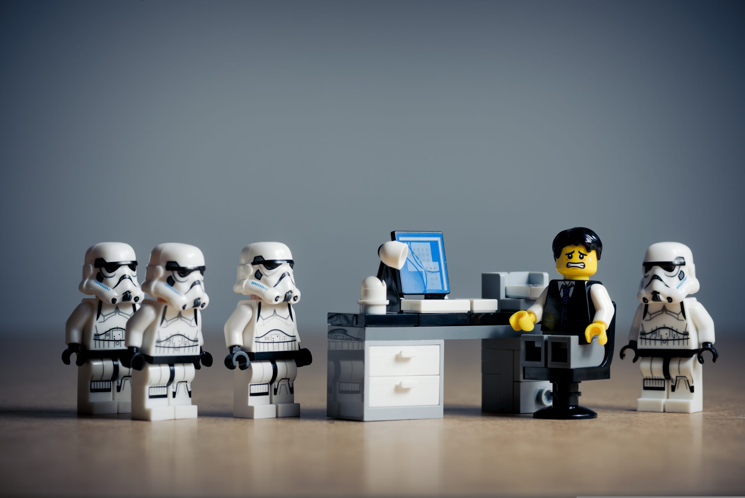 A LEGO figure sitting at a work desk made of LEGO with four Stormtrooper LEGOs surrounding him.