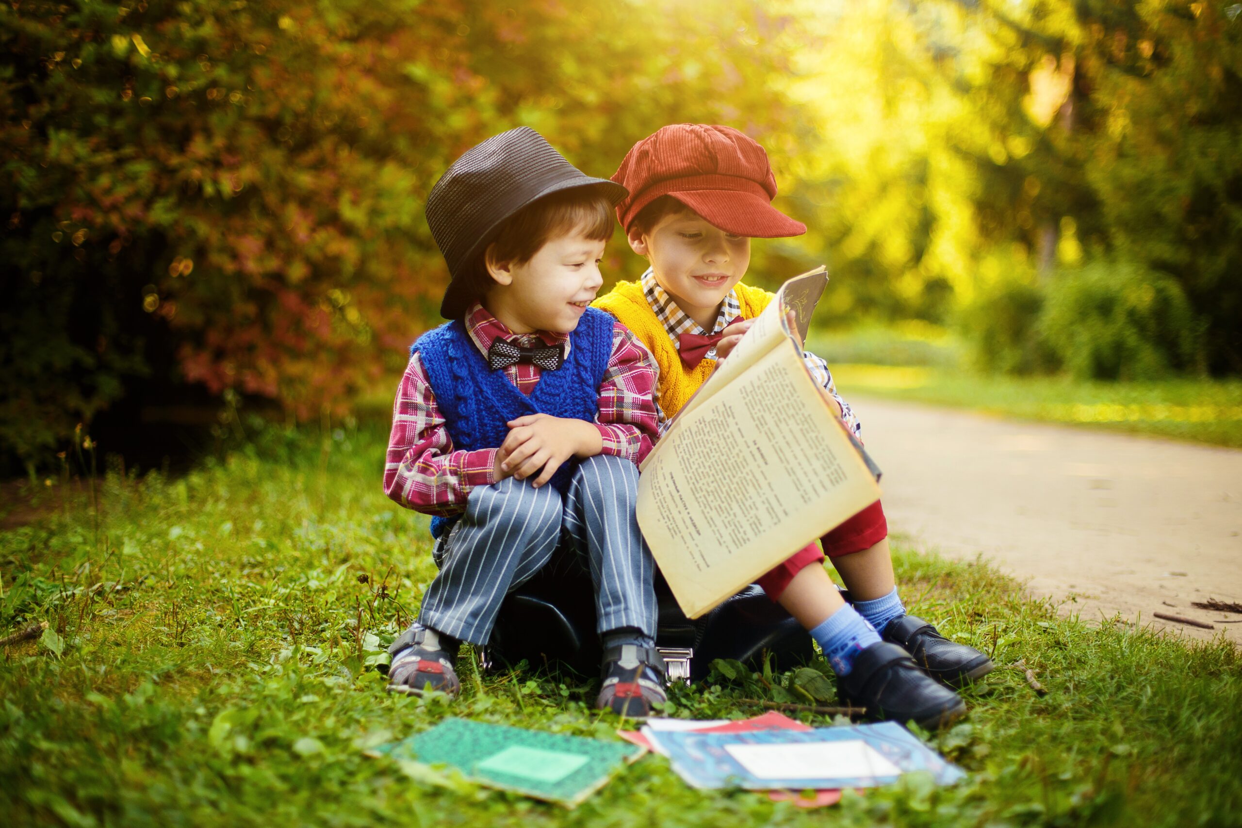 Two young boys are sitting together in the grass looking at books.